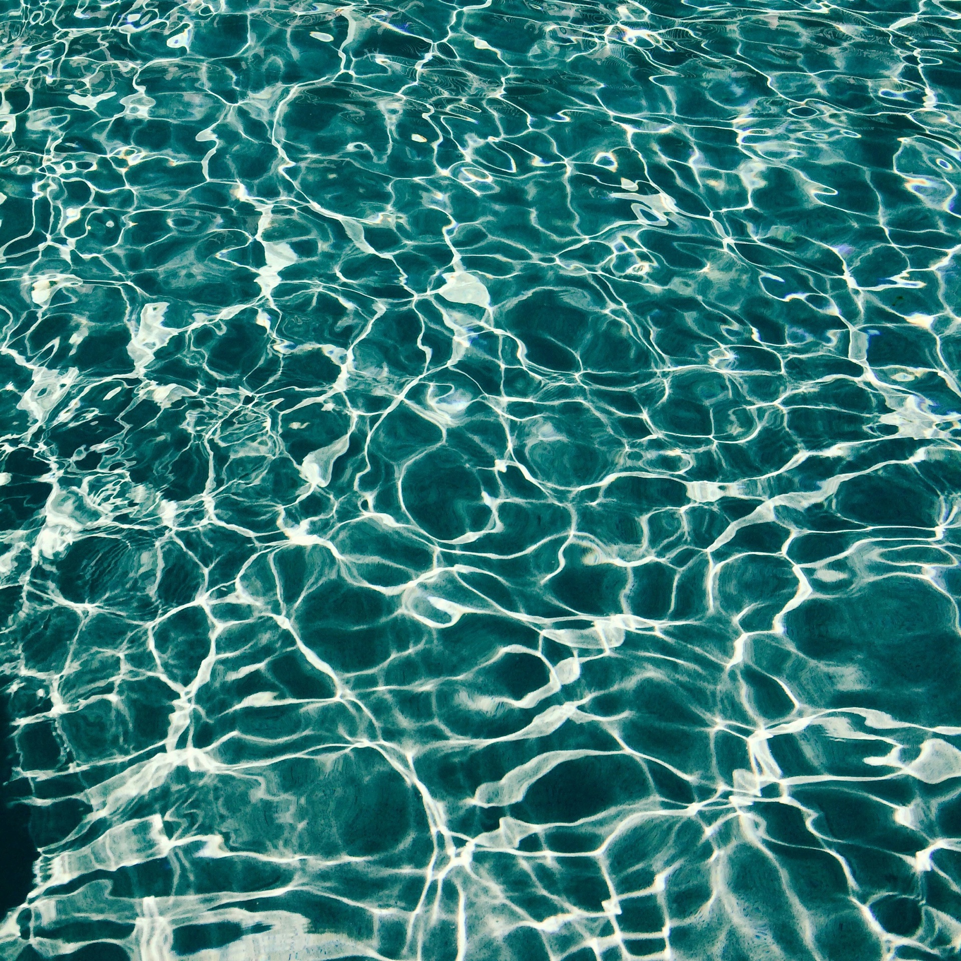 A top-down view of a modern luxury swimming pool with incredibly clear, bright blue water. A minimalist white pool skimmer net is visible in the frame, suggesting ongoing pristine maintenance. The pool edge features clean architectural lines and light-colored stone. The mood is calm and highly professional.
